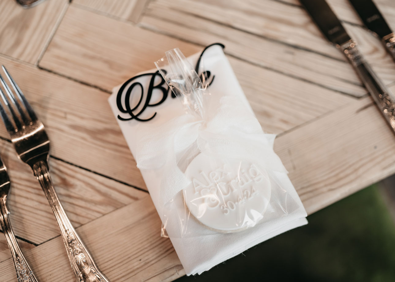 A wooden table setting with a white napkin, a round wrapped cookie, and the scripted word “Bride” placed on the napkin. Silverware is arranged on either side of the napkin.