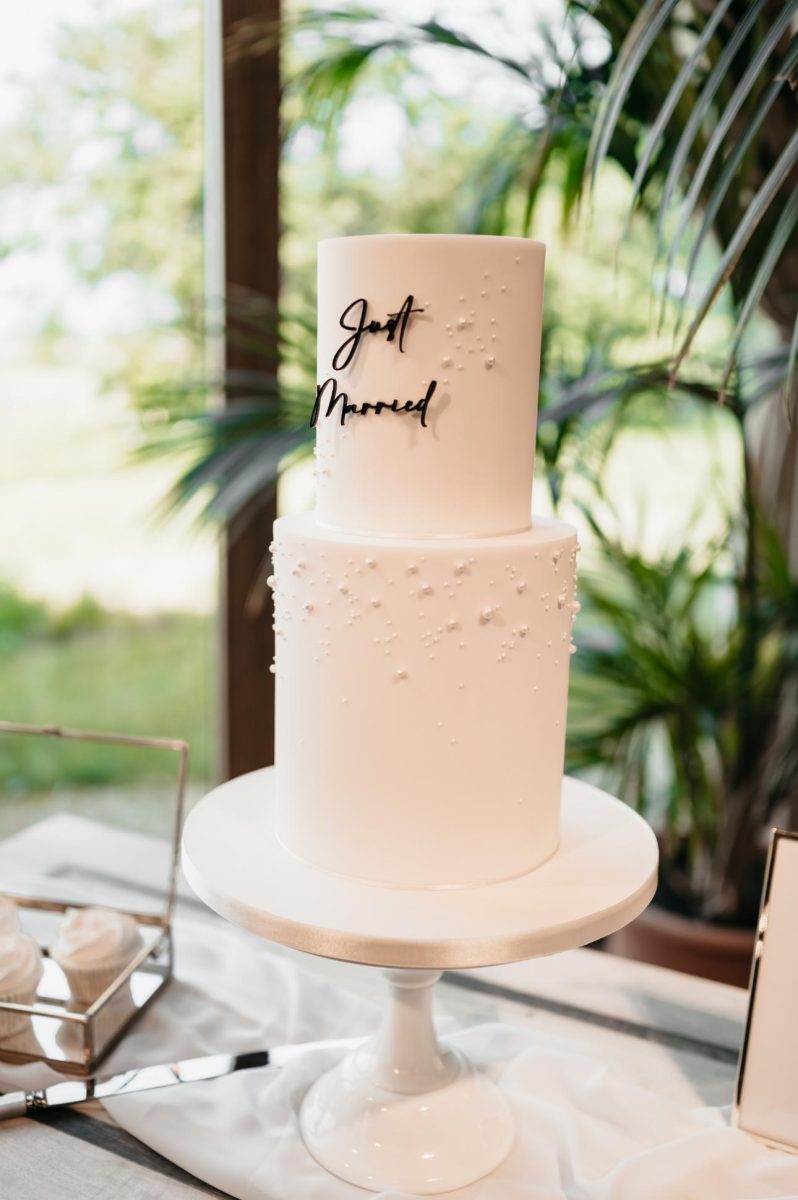 A tall, two-tier white wedding cake with "Just Married" written in black script is displayed on a pedestal stand. The cake is decorated with small white dots and is set in front of a window with greenery.