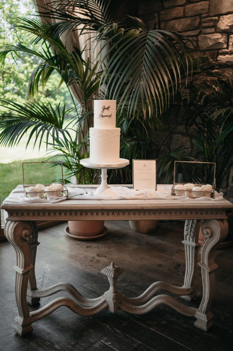 A small round white wedding cake with "Just Married" on top sits on a stand on an ornate table, flanked by greenery. Cupcakes and a menu card are also on the table.