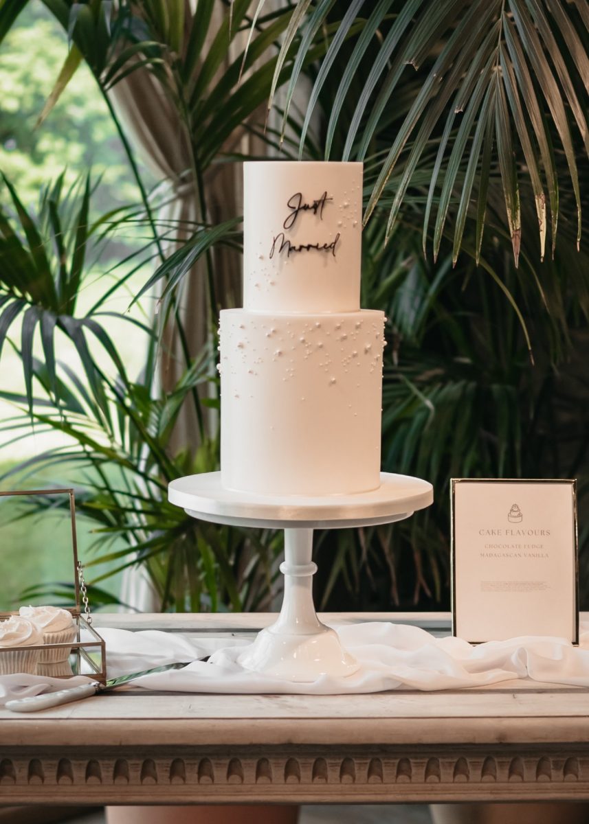 Bespoke wedding cake with two-tiers in white with "Just Married" on top, displayed on a white stand, surrounded by a leafy green background and a menu card.