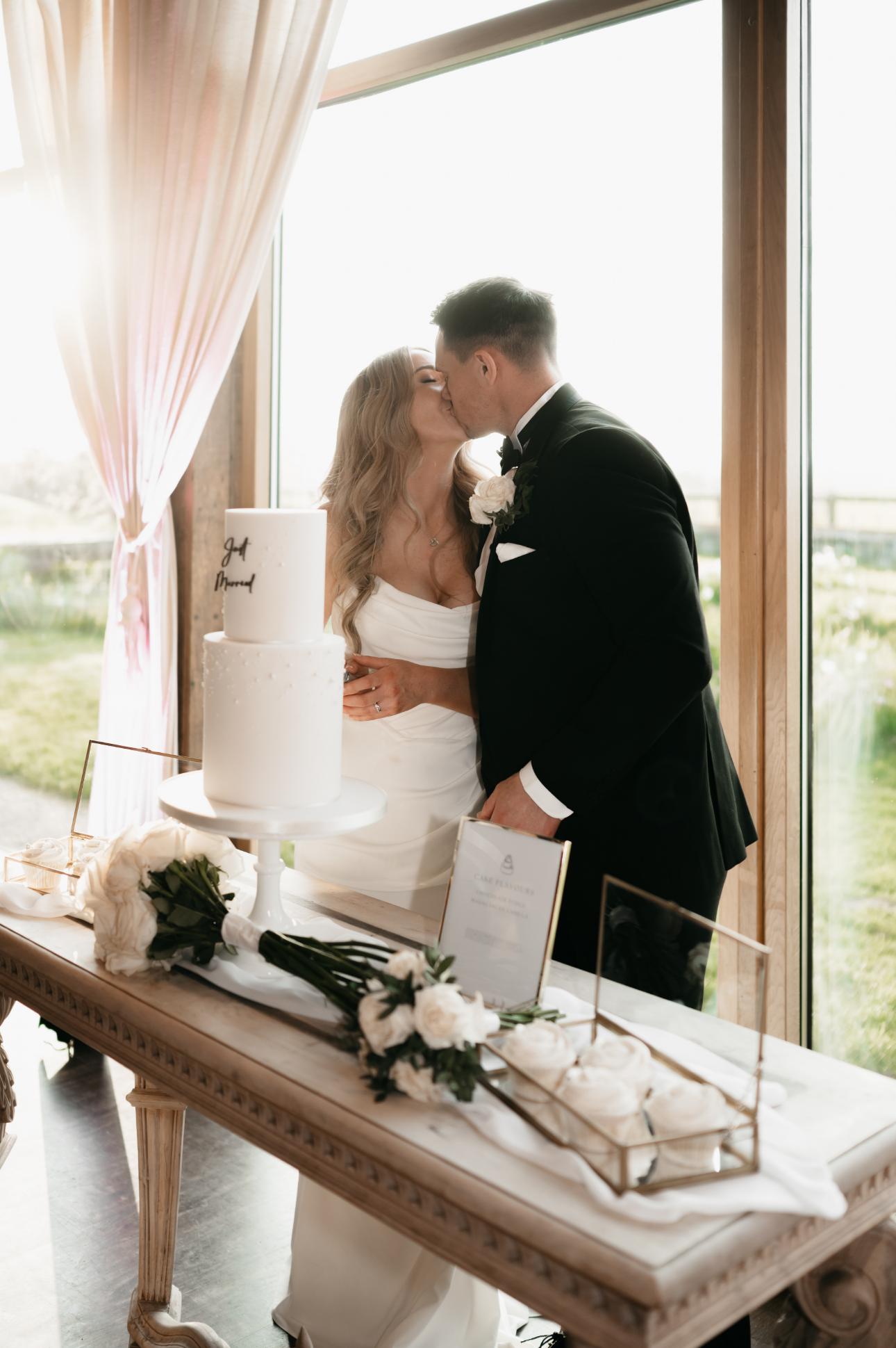 Handcrafted wedding cakes. A bride and groom share a kiss next to a white tiered wedding cake on a table. The table is decorated with white flowers and framed pictures. Sunlight streams in from large windows behind them.