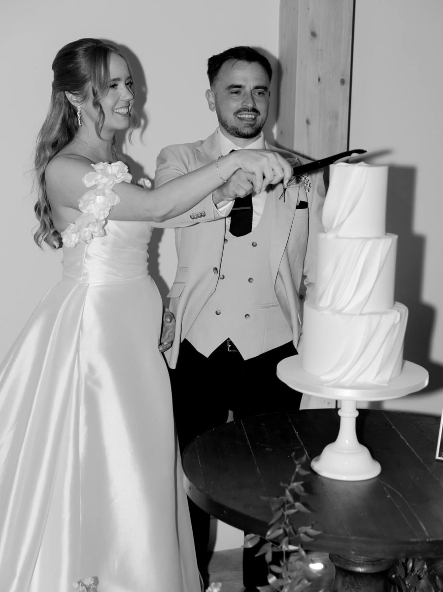 A couple in formal attire smiles while cutting a luxury fondant wedding cake at an event. The elegant, three-tiered cake sits on a round table, perfectly captured in black and white.