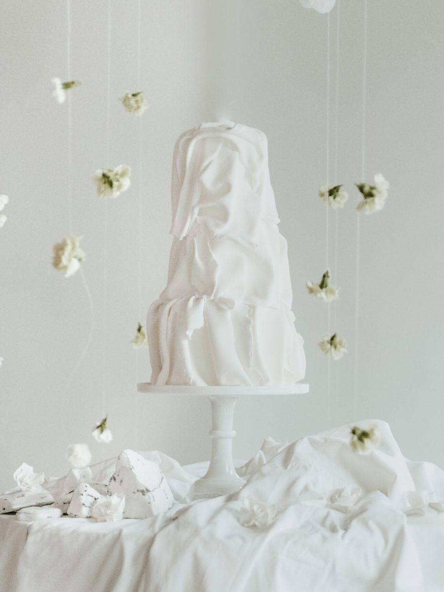 A stunning creation by a renowned cake designer from the North East, this three-tiered white cake with an abstract draped design sits elegantly on a pedestal. Delicate white flowers are artfully suspended around it.
