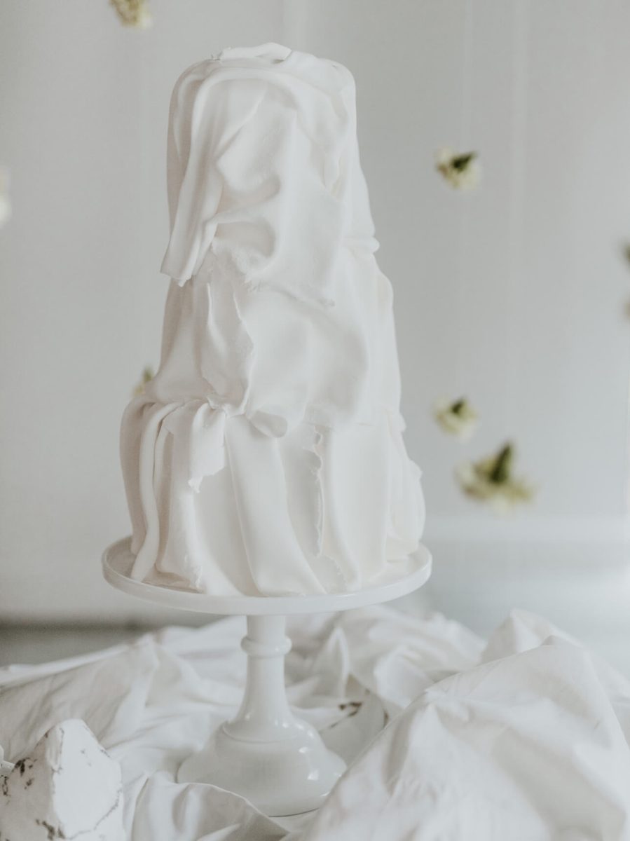 A tall, uniquely shaped white cake with a draped appearance sits elegantly on a cake stand, surrounded by white fabric and hanging flowers—a testament to the creativity of a skilled cake designer in the North East.