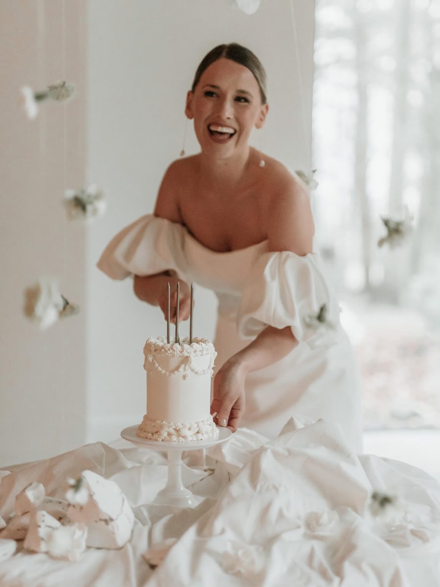 A woman in a white dress smiles broadly. She holds a beautifully decorated white cake with three candles on a stand, surrounded by elegant flowers.