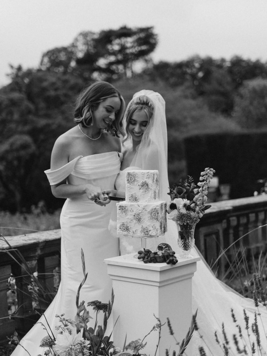 Two brides in elegant wedding dresses cut into a luxury fondant wedding cake outdoors. One wears a veil as they stand behind a pedestal adorned with flowers, surrounded by lush greenery.