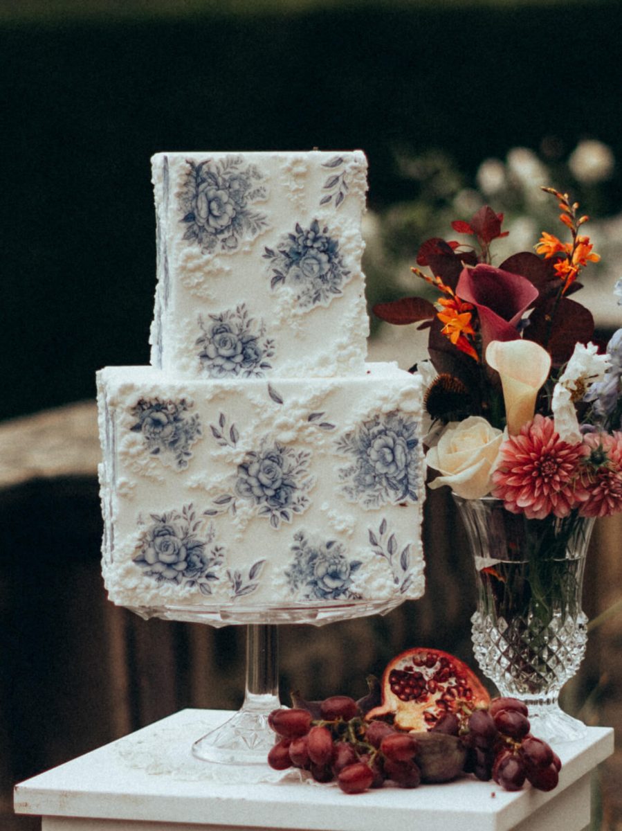 A luxury fondant wedding cake, elegantly two-tiered and white with blue floral designs, sits on a glass stand. It's beautifully accompanied by a vase of flowers, fresh grapes, and a halved pomegranate for an exquisite presentation.