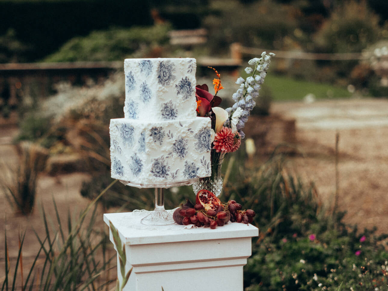 A luxury fondant wedding cake with a blue floral pattern graces a white pedestal, embellished with vibrant red and purple flowers and a pomegranate, set against an enchanting outdoor backdrop.