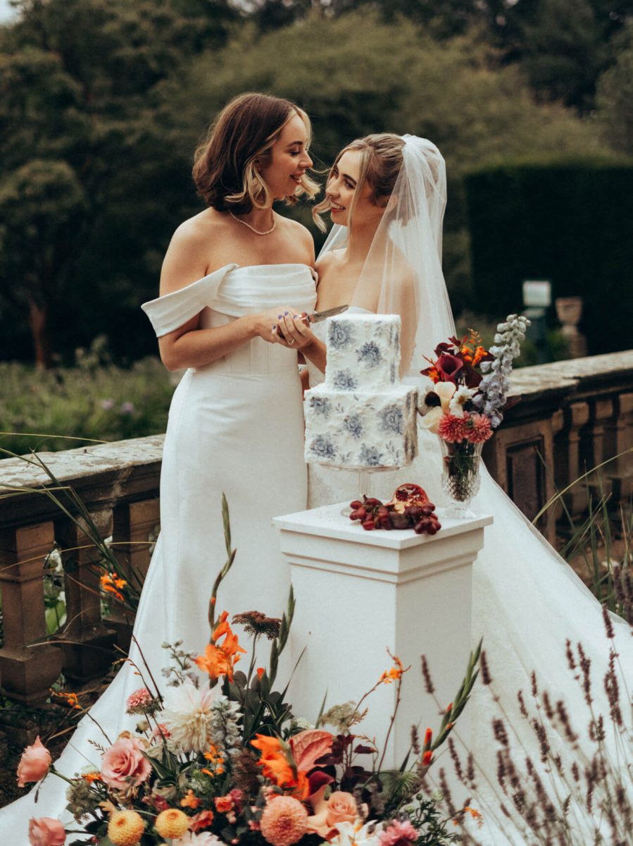 Two brides in wedding attire stand close together outdoors, smiling at each other beside a luxurious two-tiered cake decorated with fondant blue designs. Flowers are elegantly arranged around the base.