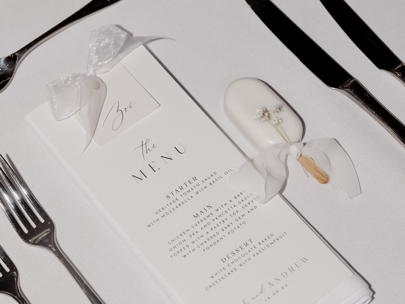 An elegant table setting showcases a menu and a decorated white oval object on a napkin, reminiscent of a luxury fondant wedding cake, flanked by shiny silverware on a pristine white tablecloth.