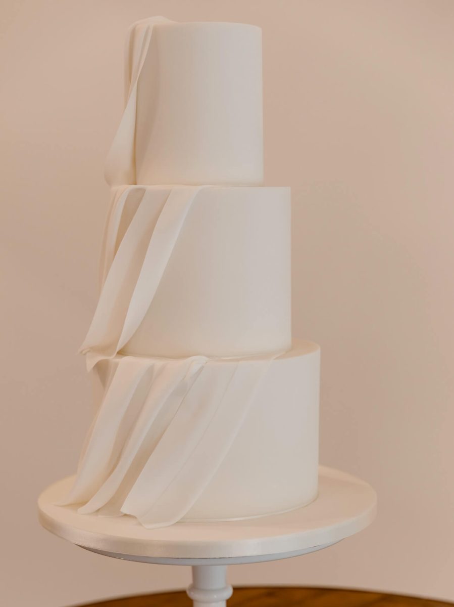 A luxurious three-tier white wedding cake with a minimalist design, featuring smooth fondant and elegant ribbon-like details, gracefully placed on a pristine white cake stand.