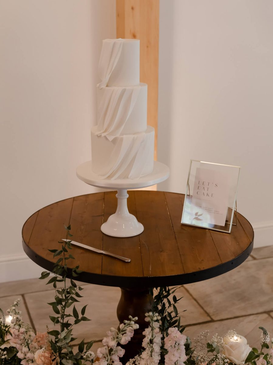 A luxury fondant wedding cake with three tiers and draped icing sits elegantly on a round wooden table, next to a small sign and cake knife, surrounded by exquisite floral decorations.