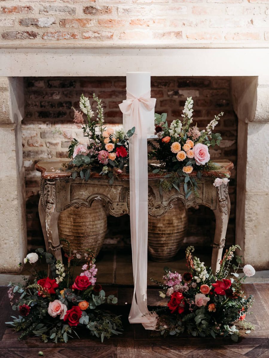 A decorative setup with a tall white candle, pink ribbon, colorful floral arrangements, and a rustic stone fireplace backdrop is perfectly complemented by a luxury fondant wedding cake.