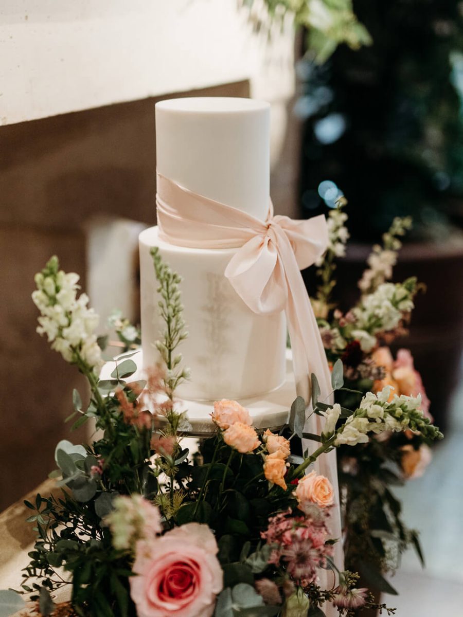 A luxurious two-tiered white cake adorned with a pink ribbon, surrounded by delicate pink and white flowers and lush greenery, creates an elegant fondant wedding centerpiece.