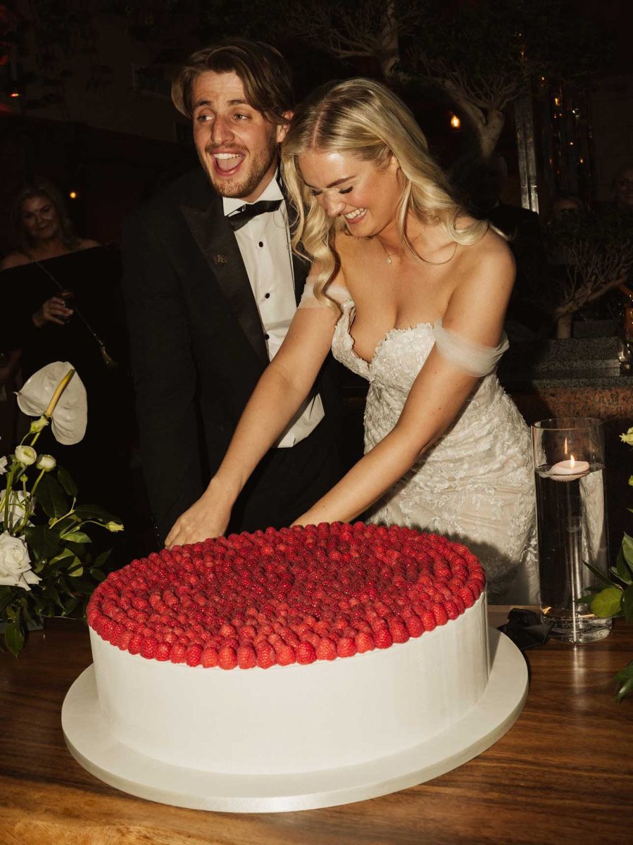 A bride and groom, both smiling, cut into a large round cake topped with raspberries at their wedding reception.