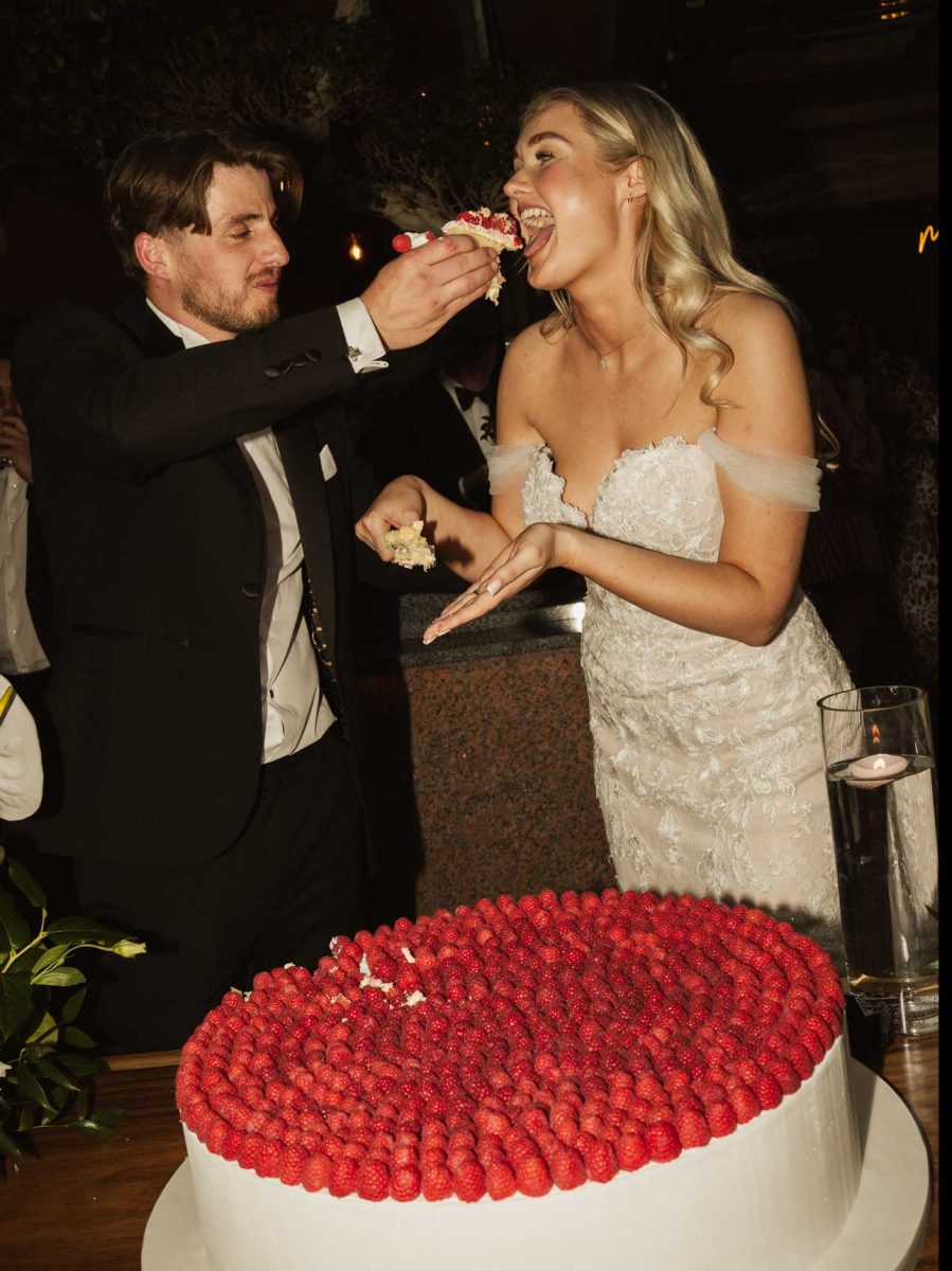 A man in a suit feeds cake to a woman in a white wedding dress next to a large cake topped with raspberries.