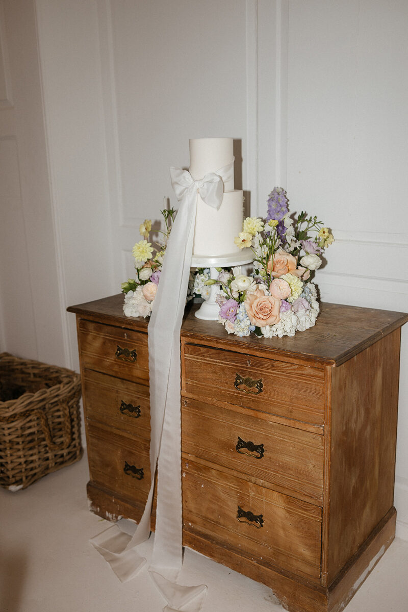 An elegant two tiered all white wedding cake with oversized ribbon bow. Set within a floral meadow provided by Rooted Up North florist.