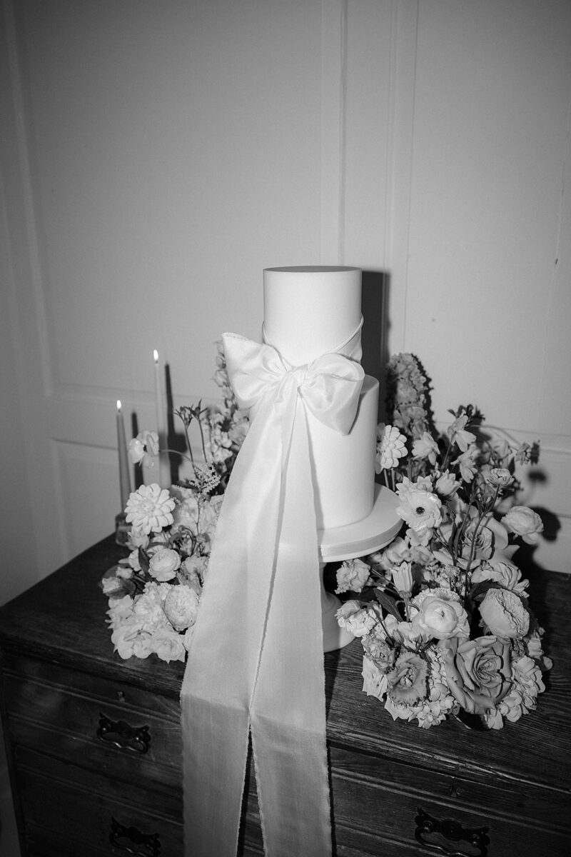 An elegant two tiered all white wedding cake with oversized ribbon bow. Set within a floral meadow provided by Rooted Up North florist.