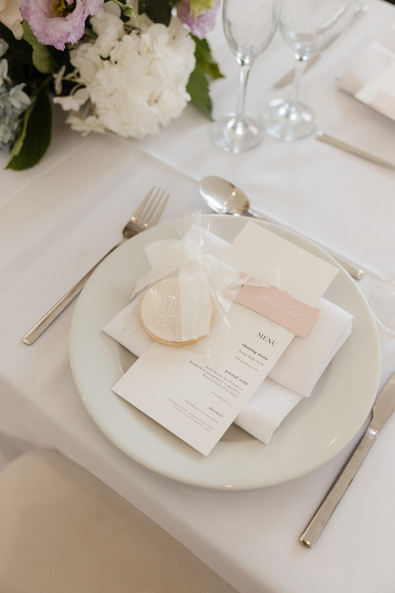 A table setting with a menu, wrapped cookie, napkin, and cutlery on a wooden table. Glassware and a name card are arranged above the plate. Created by The Little Cakery, a wedding Cake Designer North East England.