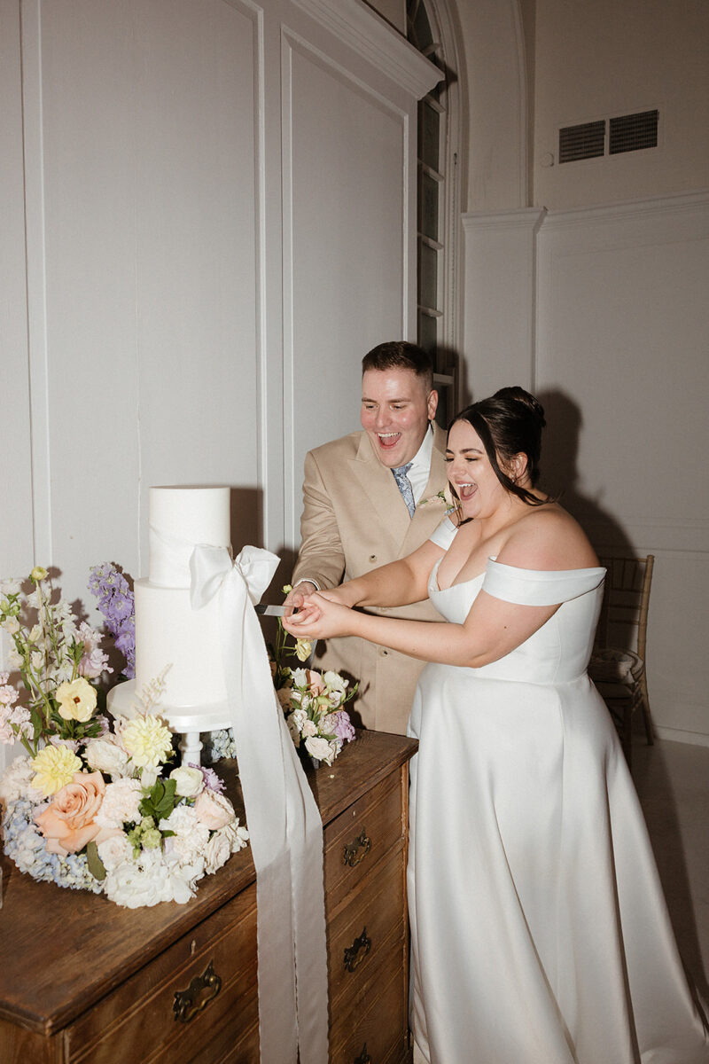 A couple in formal attire is cutting a tiered white cake decorated with flowers at a wedding reception. They are smiling and standing close to each other.