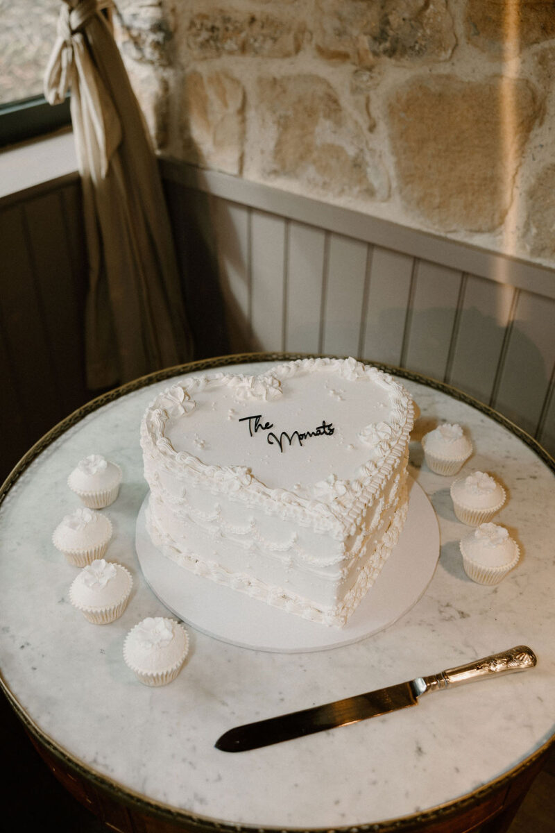 A white heart shaped wedding cake with vintage lambeth piped details, pearls and an acrylic charm. Set up with floral wedding cupcakes.