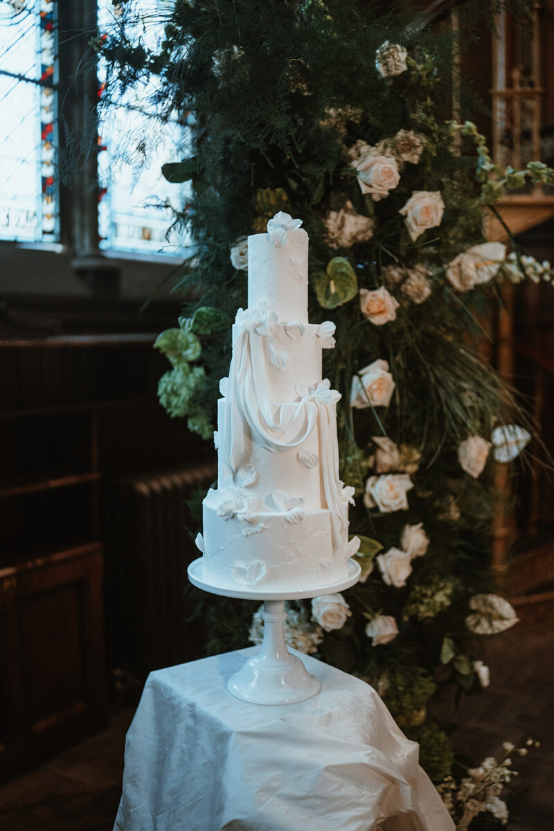 A four tiered all white statement wedding cake with textured fondant, fondant drapes and handmade sugar petals. Set up at The Common Room Newcastle Upon Tyne City Centre Wedding. With Sunday Girl Florist and Emma Jordan Weddings.