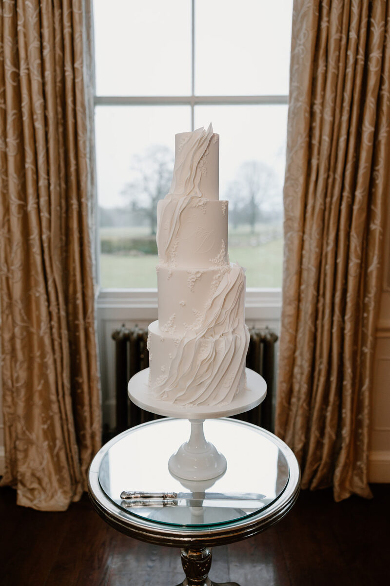 A statement all white wedding cake with textured details and couples initials. Set up in the backdrop of a grand wedding venue.