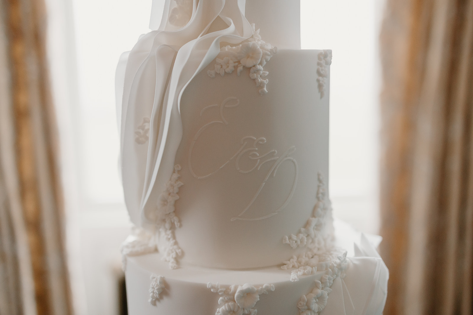 A statement all white wedding cake with textured details and couples initials. Set up in the backdrop of a grand wedding venue.